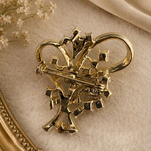 May include: A gold-toned brooch with an ornate design, featuring a floral or crown-like motif. The back of the brooch reveals a pin clasp for secure attachment. The brooch is set against a neutral background, highlighting its intricate details.