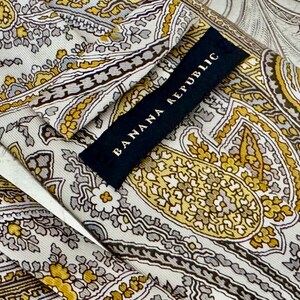 May include: Close-up of a silk tie with a paisley pattern in shades of gold, gray, and cream. The tie features the "Banana Republic" label in black. The intricate design is visible throughout the fabric.