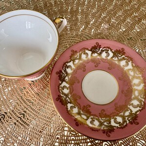 May include: A vintage teacup and saucer set. The teacup is white inside with a pink exterior and gold trim. The saucer is pink with a white center and gold filigree design. The set is on a gold-colored woven mat.