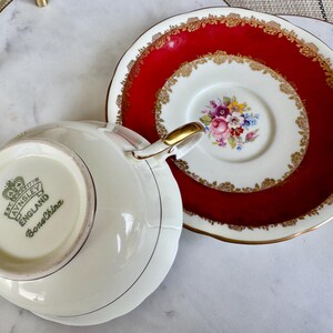 May include: A white teacup and saucer with a red and gold floral design. The teacup has a gold rim and the saucer has a red rim with gold accents. The teacup has the words "Aynsley England Bone China" on the bottom.