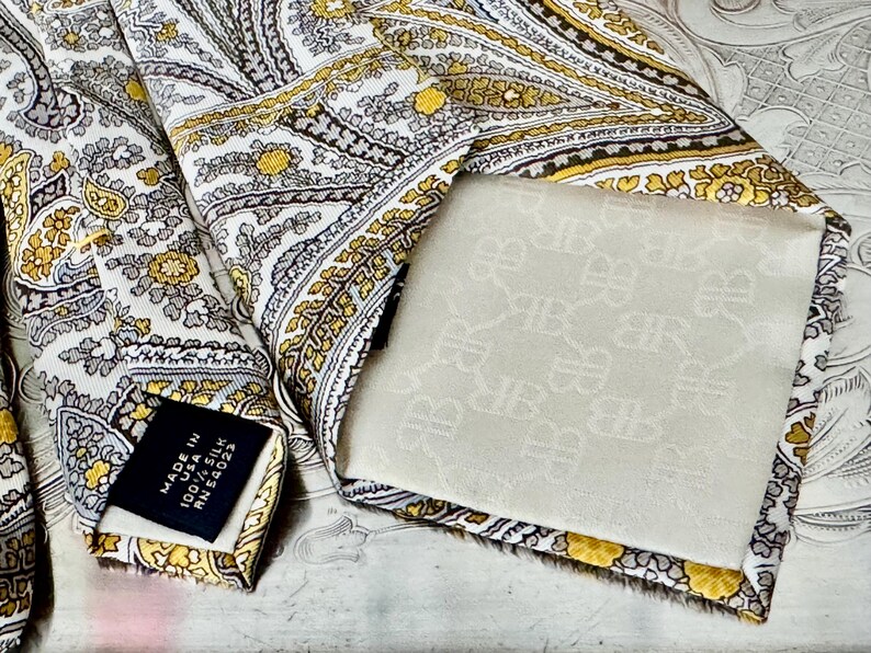 May include: A silk tie with a paisley pattern in white, gold, and gray. The tie has a label that reads "Made in USA 100% Silk". The back of the tie is a cream color with a repeating pattern.