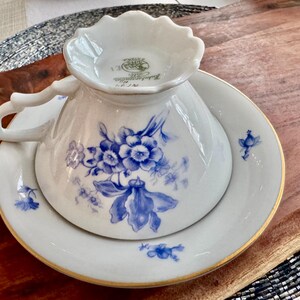 May include: A white porcelain teacup and saucer set with a blue floral pattern. The cup has a scalloped rim and a gold trim. The saucer has a gold rim and a blue floral pattern.
