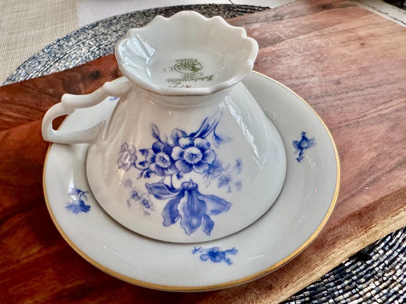 May include: A white porcelain teacup and saucer set with a blue floral pattern. The cup has a scalloped rim and a gold trim. The saucer has a gold rim and a blue floral pattern.