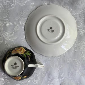Pode incluir: Um conjunto de ch&aacute;vena e pires de porcelana Royal Albert visto pela parte inferior. A ch&aacute;vena &eacute; preta com padr&otilde;es de rosas amarelas e uma asa branca. Ambas as pe&ccedil;as apresentam o selo da coroa Royal Albert England nas bases brancas.