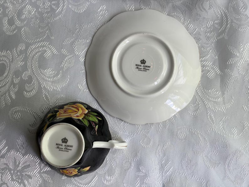Pode incluir: Um conjunto de ch&aacute;vena e pires de porcelana Royal Albert visto pela parte inferior. A ch&aacute;vena &eacute; preta com padr&otilde;es de rosas amarelas e uma asa branca. Ambas as pe&ccedil;as apresentam o selo da coroa Royal Albert England nas bases brancas.