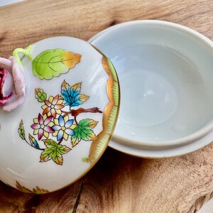 May include: A white porcelain trinket box with a floral design. The lid features a 3D rose with pink and red petals, green leaves, and a butterfly. The box has a gold trim around the edge.