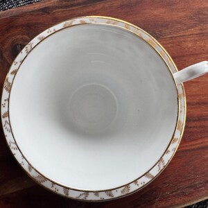May include: A white teacup with a gold rim and a delicate floral pattern. The cup is empty and sitting on a wooden surface.