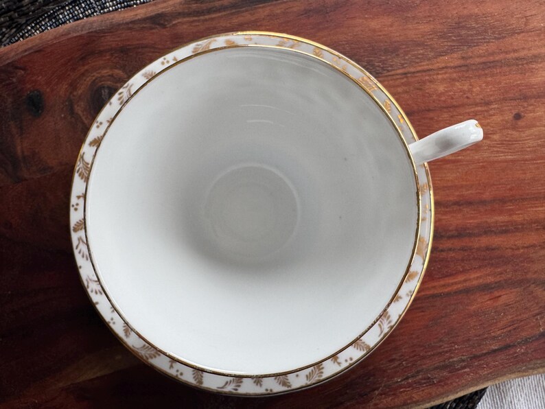 May include: A white teacup with a gold rim and a delicate floral pattern. The cup is empty and sitting on a wooden surface.