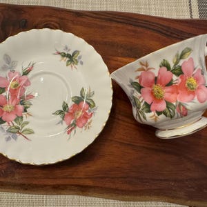 Puede incluir: Un juego de taza y platillo de t&eacute; blanco con un dise&ntilde;o floral con rosas rosas y hojas verdes. El juego tiene un borde dorado.