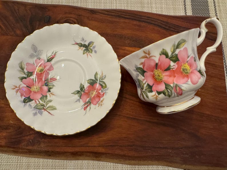Puede incluir: Un juego de taza y platillo de t&eacute; blanco con un dise&ntilde;o floral con rosas rosas y hojas verdes. El juego tiene un borde dorado.