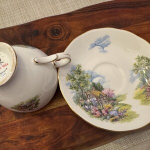 May include: A white teacup and saucer set with a gold rim. The saucer features a painted scene of a cottage in a garden with trees and flowers. The teacup has the words "Royal Vale" printed on the bottom.