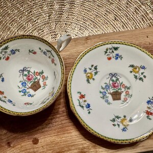 Pu&ograve; includere: Una tazza da t&egrave; e un piattino bianchi con un bordo dorato e un motivo floreale. Il motivo presenta fiori rosa, gialli e blu e foglie verdi. La tazza e il piattino hanno un cestino di fiori al centro.