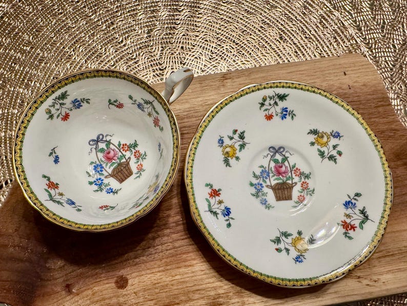 Pu&ograve; includere: Una tazza da t&egrave; e un piattino bianchi con un bordo dorato e un motivo floreale. Il motivo presenta fiori rosa, gialli e blu e foglie verdi. La tazza e il piattino hanno un cestino di fiori al centro.