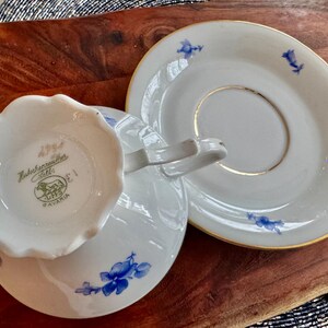 May include: A white teacup and saucer with a blue floral pattern. The teacup has a scalloped edge and the saucer has a gold rim. The bottom of the teacup has the text "Hutschenreuther 1794 Selb Bavaria" in gold lettering.