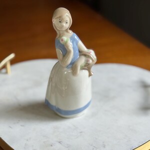 K&ouml;nnte beinhalten: Porzellanfigur eines sitzenden M&auml;dchens, gekleidet in ein blaues Oberteil und einen wei&szlig;en Rock mit blauem Streifen. Sie h&auml;lt ein gr&uuml;nes und rosa Objekt. Die Figur steht auf einem wei&szlig;en Marmortablett mit Goldrand.