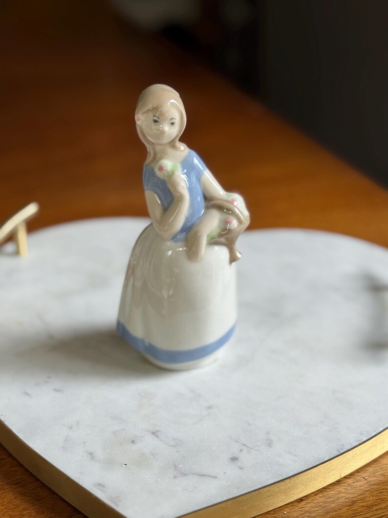 K&ouml;nnte beinhalten: Porzellanfigur eines sitzenden M&auml;dchens, gekleidet in ein blaues Oberteil und einen wei&szlig;en Rock mit blauem Streifen. Sie h&auml;lt ein gr&uuml;nes und rosa Objekt. Die Figur steht auf einem wei&szlig;en Marmortablett mit Goldrand.