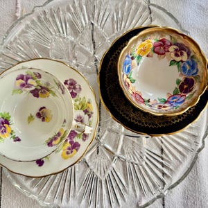 以下が含まれることがあります： 花柄のヴィンテージティーカップとソーサー2客。紫と黄色のパンジーが描かれたものと、カラフルな花柄のものがあります。どちらも金色の縁取り。透明なガラスのサービングプレートの上に置かれています。