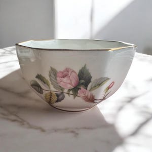 Pode incluir: Uma tigela de porcelana branca com borda dourada e design floral. A tigela apresenta uma rosa rosa com folhas verdes e caules marrons. A tigela tem uma forma octogonal &uacute;nica.