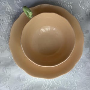 May include: A peach-colored ceramic teacup and matching saucer set. The teacup features a smooth, rounded bowl with a scalloped rim and a pale green handle. The saucer has a matching scalloped edge, creating a cohesive look for this drinkware.