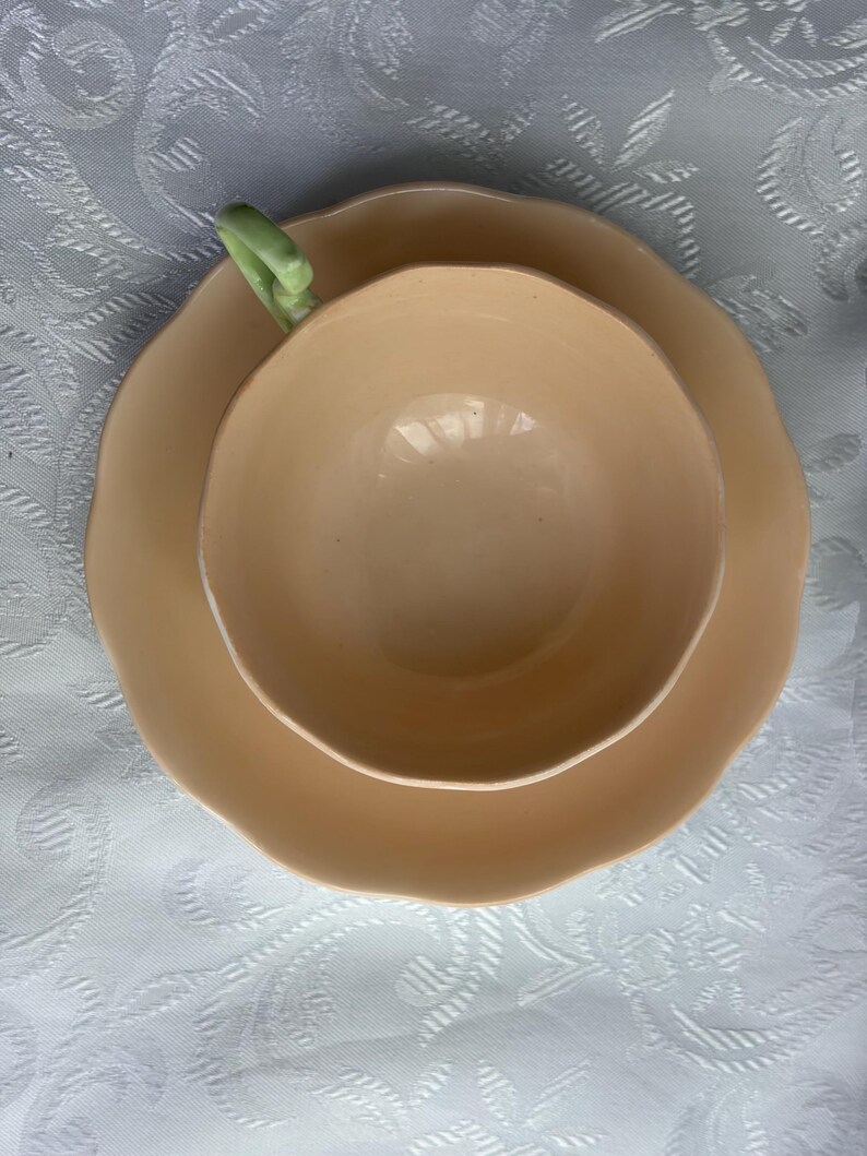 May include: A peach-colored ceramic teacup and matching saucer set. The teacup features a smooth, rounded bowl with a scalloped rim and a pale green handle. The saucer has a matching scalloped edge, creating a cohesive look for this drinkware.
