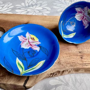 Puede incluir: Un juego de taza y platillo de t&eacute; azul y blanco con un dise&ntilde;o floral. La taza y el platillo est&aacute;n decorados con una flor rosa y amarilla con hojas verdes. El fondo es de color azul s&oacute;lido.