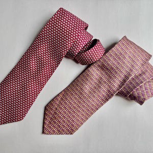 Puede incluir: Se muestran dos corbatas. La corbata de la izquierda es de color rojo intenso con un patr&oacute;n geom&eacute;trico blanco repetitivo. La corbata de la derecha es de un color malva apagado con un patr&oacute;n de peque&ntilde;as formas verticales claras y abstractas.