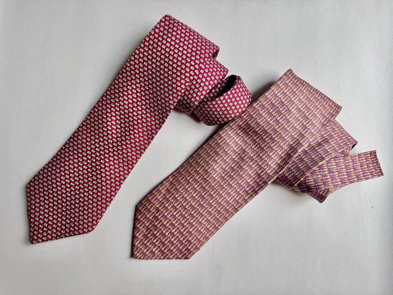 Puede incluir: Se muestran dos corbatas. La corbata de la izquierda es de color rojo intenso con un patr&oacute;n geom&eacute;trico blanco repetitivo. La corbata de la derecha es de un color malva apagado con un patr&oacute;n de peque&ntilde;as formas verticales claras y abstractas.
