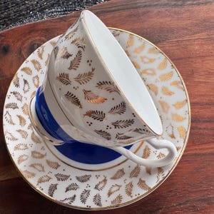 May include: A white teacup and saucer with a gold and white leaf pattern. The saucer has a blue band around the edge.