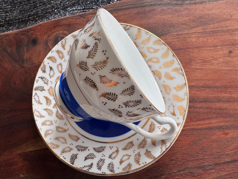 May include: A white teacup and saucer with a gold and white leaf pattern. The saucer has a blue band around the edge.