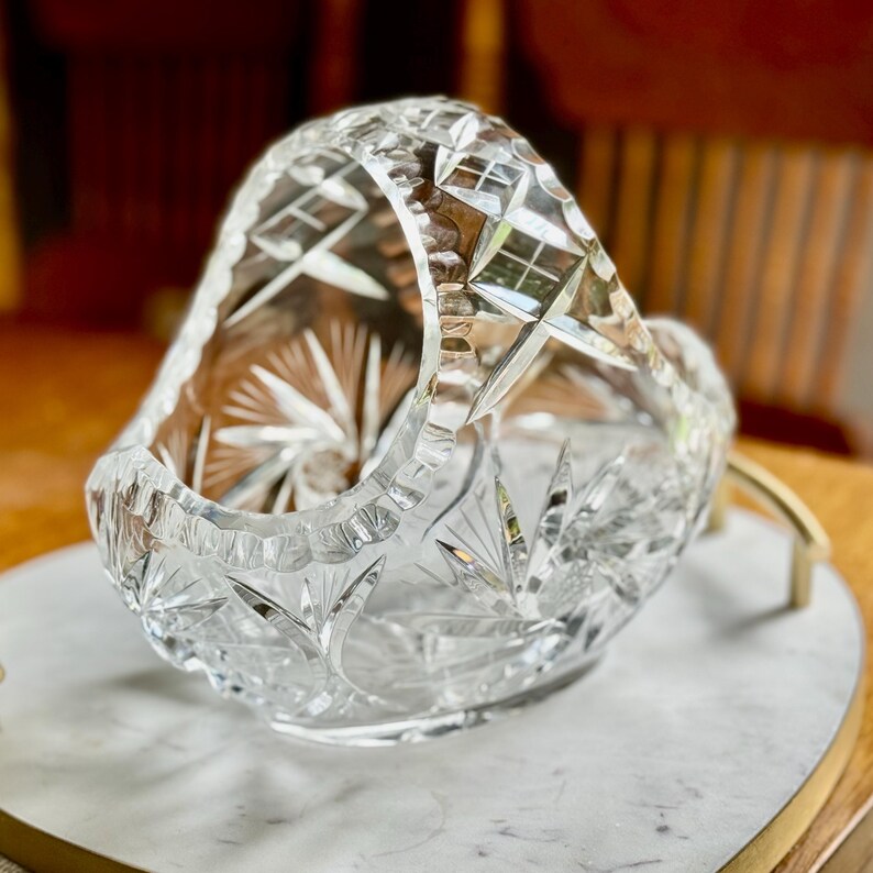 May include: A clear, cut-glass basket with a handle. The basket features intricate starburst and geometric patterns. The handle is also cut with a diamond pattern. The basket is sitting on a white and gray marble tray.