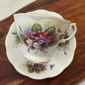 May include: A white teacup and saucer set with a floral design featuring purple and pink violets. The cup has a scalloped edge and a gold trim.