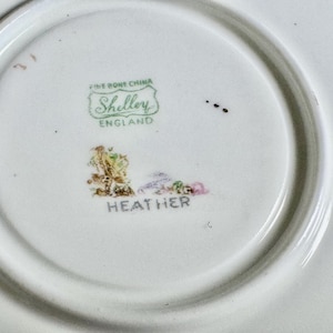 May include: The underside of a white fine bone china saucer featuring a green Shelley England maker's mark. Below the mark is a small, colorful floral illustration and the word Heather printed in black capital letters on the ceramic surface.