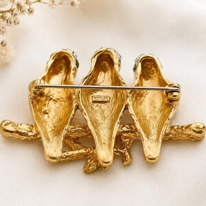 May include: A gold-toned brooch featuring three birds perched on a branch. The birds have black accents on their heads and are detailed with textured feathers. The brooch has a pin closure on the back and is marked with the word "BUTLER".