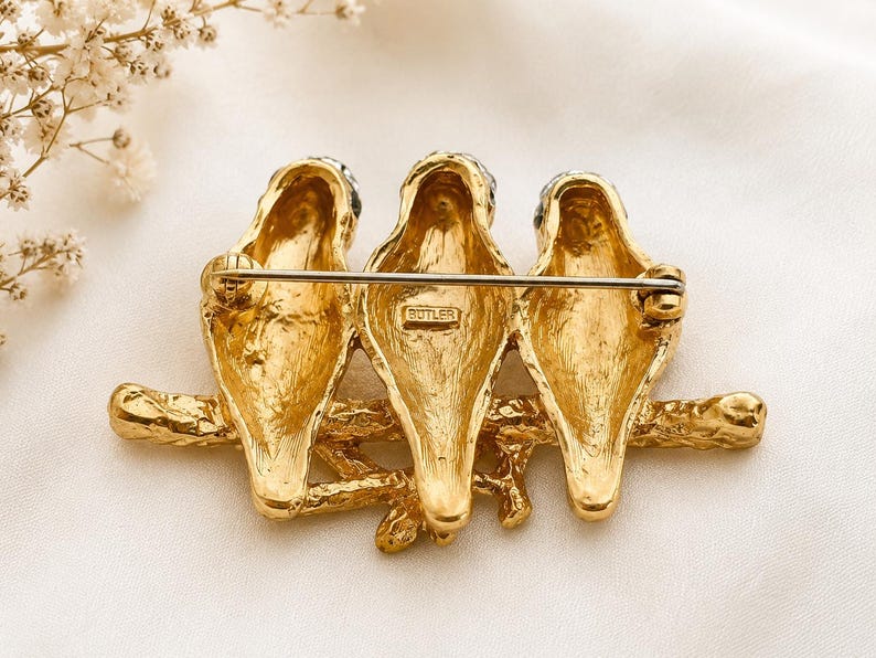 May include: A gold-toned brooch featuring three birds perched on a branch. The birds have black accents on their heads and are detailed with textured feathers. The brooch has a pin closure on the back and is marked with the word "BUTLER".