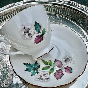 May include: White teacup and saucer set with floral and leaf design in pink, green, and purple. The teacup and saucer have gold trim and are on a silver tray. A classic tea set.