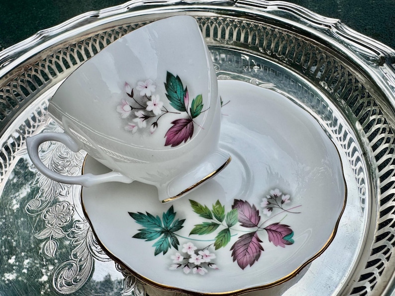 May include: White teacup and saucer set with floral and leaf design in pink, green, and purple. The teacup and saucer have gold trim and are on a silver tray. A classic tea set.