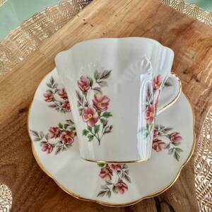 May include: A white teacup and saucer with a floral pattern. The cup has a scalloped rim and a gold trim. The saucer has a matching floral pattern and a gold trim.