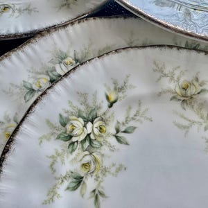 May include: Set of three white ceramic plates with a gold rim and a floral design featuring white roses and green leaves.