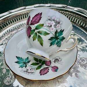 May include: White teacup and saucer set with a floral design featuring pink, green, and white flowers and leaves. The cup and saucer have gold trim. The set is resting on a silver tray.
