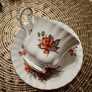 Pode incluir: Uma ch&aacute;vena de ch&aacute; e um pires brancos com um desenho floral que apresenta uma rosa vermelha grande e rosas cor-de-rosa menores. A ch&aacute;vena e o pires t&ecirc;m uma borda dourada.