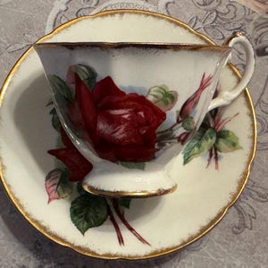 May include: A white teacup and saucer set with a red rose design. The teacup and saucer have gold trim. The teacup features a large red rose with green leaves and buds. The saucer also has a matching rose design.
