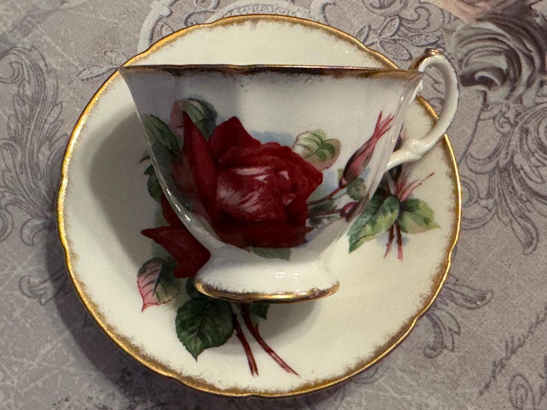 May include: A white teacup and saucer set with a red rose design. The teacup and saucer have gold trim. The teacup features a large red rose with green leaves and buds. The saucer also has a matching rose design.