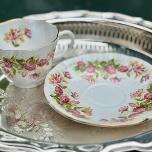 May include: A white teacup and saucer set with a floral design in pink, yellow, and green. The teacup has a gold rim and handle. The set is on a silver tray.
