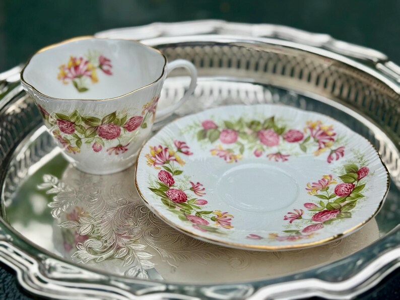 May include: A white teacup and saucer set with a floral design in pink, yellow, and green. The teacup has a gold rim and handle. The set is on a silver tray.
