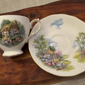 May include: A white teacup and saucer set with a cottagecore design. The teacup and saucer feature a painted scene of a cottage with a thatched roof, surrounded by flowers and trees. The background is a light blue sky with white clouds.