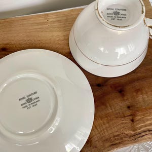 Pode incluir: Um conjunto de ch&aacute;vena e pires de porcelana Royal Stafford branca. A ch&aacute;vena e o pires s&atilde;o brancos com detalhes dourados. O log&oacute;tipo e o texto Royal Stafford est&atilde;o no pires e na base da ch&aacute;vena. Fabricado na Inglaterra em 1845.