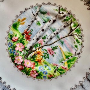 Pu&ograve; includere: Un piatto in ceramica bianca con un design decorativo di betulle e fiori. Il piatto ha un bordo ondulato con un bordo grigio.