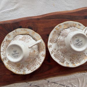 May include: Two white teacups and saucers with gold floral patterns. The saucers have a scalloped edge. The teacups have a gold trim and the words "Queen Anne" printed on the bottom.