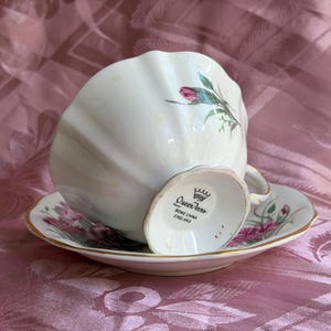 May include: A white bone china teacup and saucer set featuring a scalloped edge and a floral pattern with pink flowers and green leaves. The base of the cup is marked with a crown logo and the text Queen Anne Bone China England in black ink.