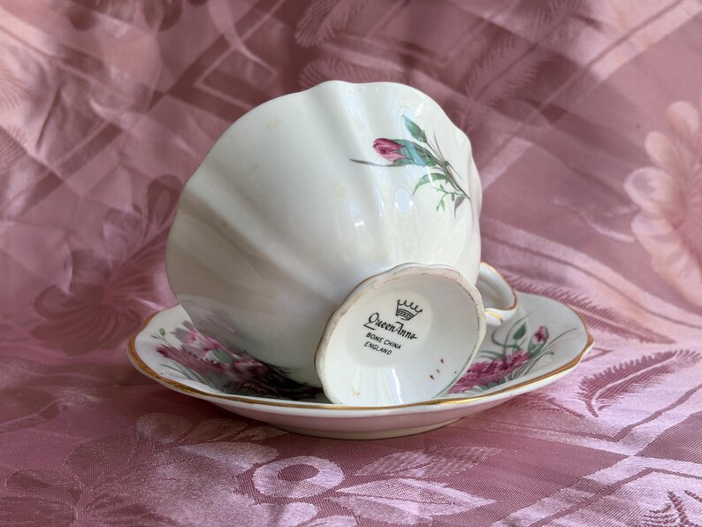 May include: A white bone china teacup and saucer set featuring a scalloped edge and a floral pattern with pink flowers and green leaves. The base of the cup is marked with a crown logo and the text Queen Anne Bone China England in black ink.